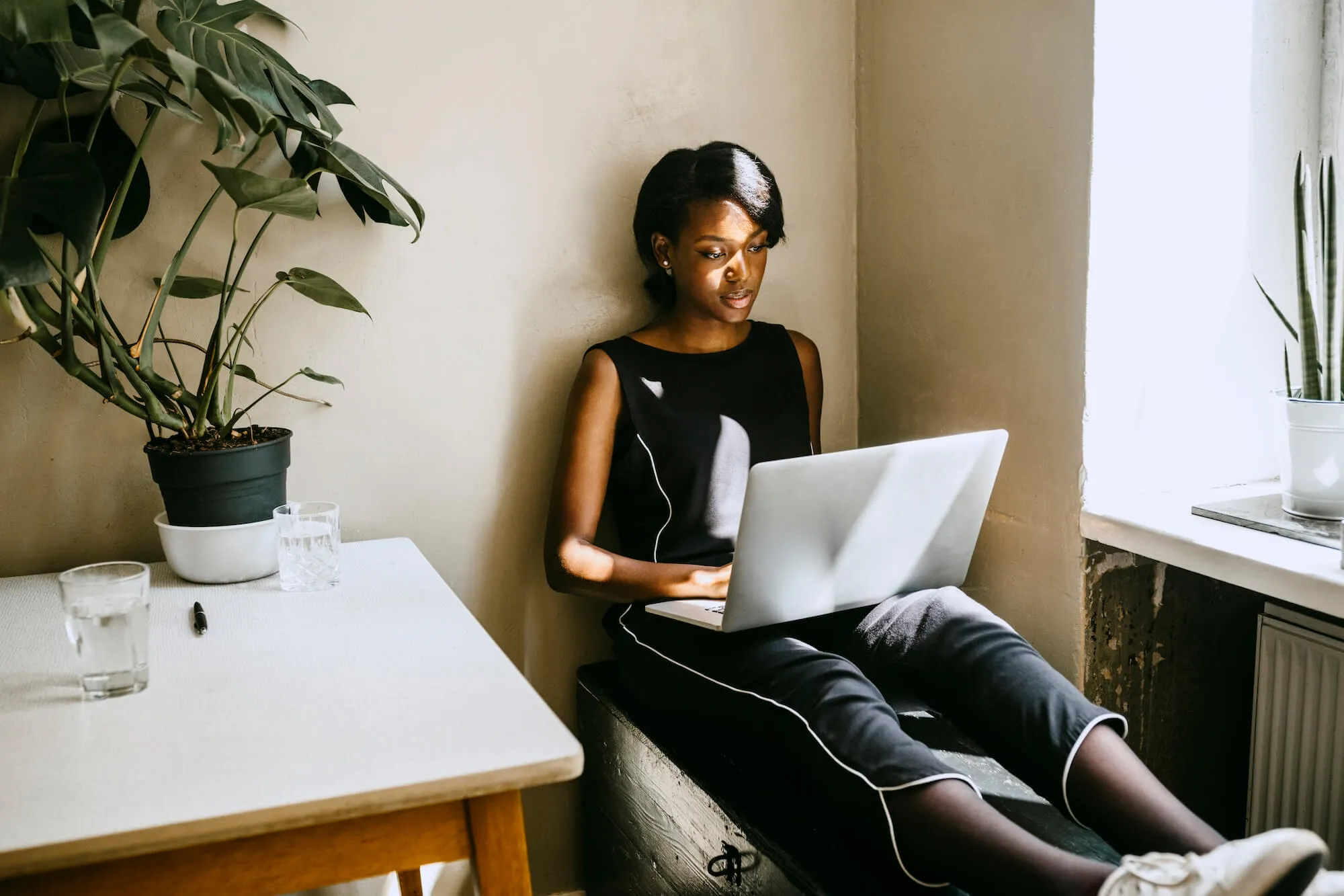 black woman sitting on a desk, with her laptop on her lap. she is wearing a black top and jeans and is next to a desk with a plant on it and a window sill